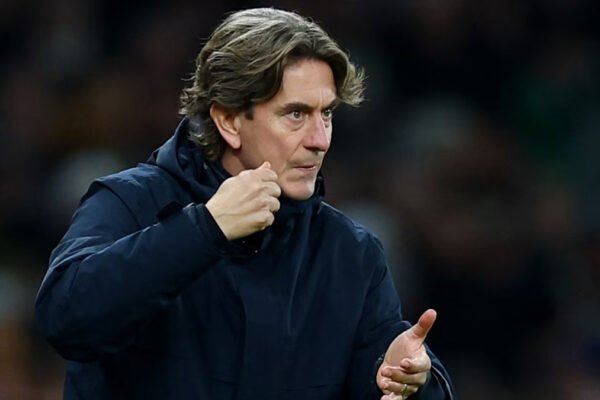 Head coach on the touchline during a night match, crowd visible behind him.