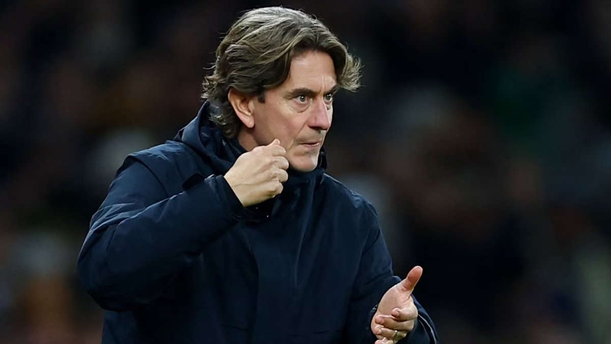 Head coach on the touchline during a night match, crowd visible behind him.